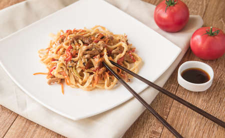 Egg udon noodles with pork, vegetables and sesame seeds on a white plate, wooden table, shinese chopsticks, close-upの写真素材