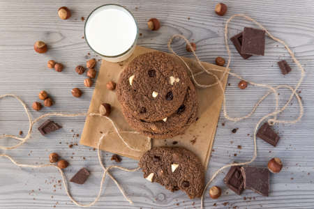 Chocolate cookies with hazelnuts, white chocolate and dark chocolate on parchment and glass of milk, wooden background, flat lay, top viewの写真素材