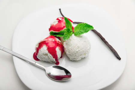 Three scoops of vanilla ice cream with strawberry syrup and mint on a white plate and a vanilla bean, white backgroundの写真素材