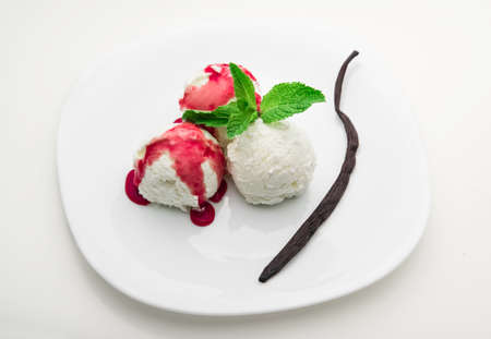 Three scoops of vanilla ice cream with strawberry syrup and mint on a white plate and a vanilla bean, white backgroundの写真素材