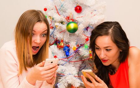Two beautiful girls lying next to a Christmas tree using a mobile phonesの写真素材