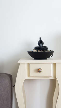 Front view of a decorative zen water fountain on top of a small white table next to a gray couch. A...