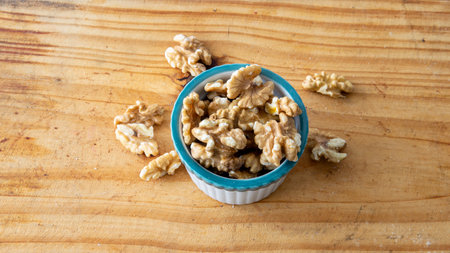 top view of a small white creamy bowl loaded with walnuts. Placed on top of a wooden table with...