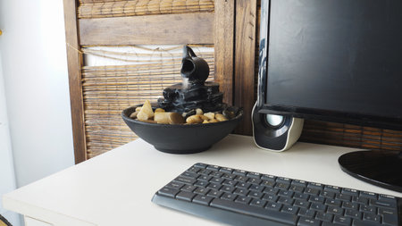 <p>Close view of a decorative zen water fountain next to a keyboard, and a computer monitor. on top of a white wooden desk</p>の写真素材