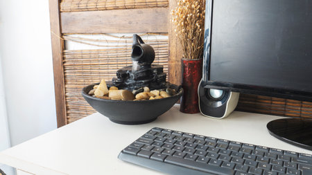 <p>Close view of a decorative zen water fountain next to a keyboard, a monitor and dry flowers decorative bouquet. on top of a white wooden desk</p>の写真素材