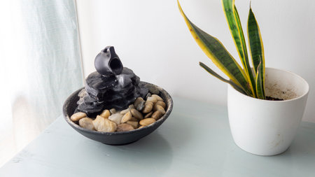 zen water fountain placed on top of a white wooden table next to a window and a green plant in a potの写真素材