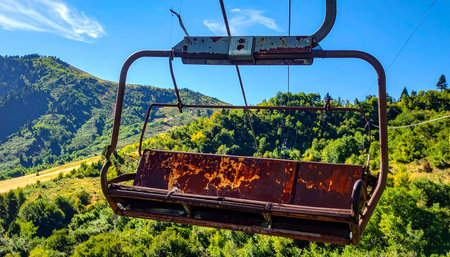 An old, rusty ski lift chair hangs silently against a backdrop of vibrant green mountains and a clear blue sky.の素材