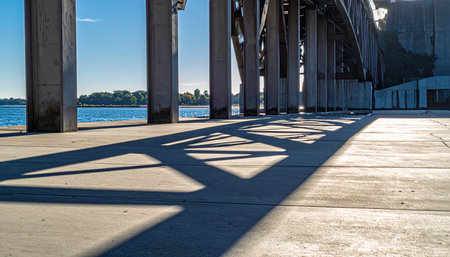 From a low perspective, the morning sun casts long, geometric shadows from an industrial bridge's steel truss onto the concrete pier below.の素材