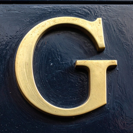 A close-up of a polished brass letter 'G' in a classic serif font, mounted on a dark, textured surface.の素材