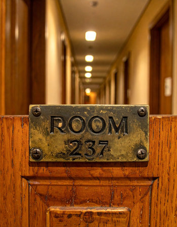 A close-up of a tarnished brass plaque for Room 237 on a wooden door. In the background, a long, dimly lit hotel hallway stretches into the distance, creating a sense of suspense and mystery.の素材
