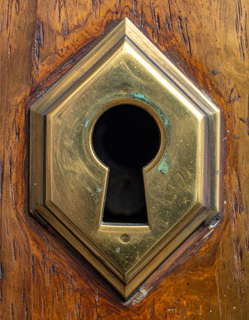 A close-up of an antique brass keyhole set in a rich, grained wood surface. This timeless detail evokes a sense of mystery, secrecy, and the potential for discovery.の素材