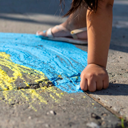 A close-up view captures the simple joy of childhood creativity. A small hand firmly grips a piece of chalk, adding vibrant blue and yellow colors to a masterpiece on the warm summer pavement.の素材