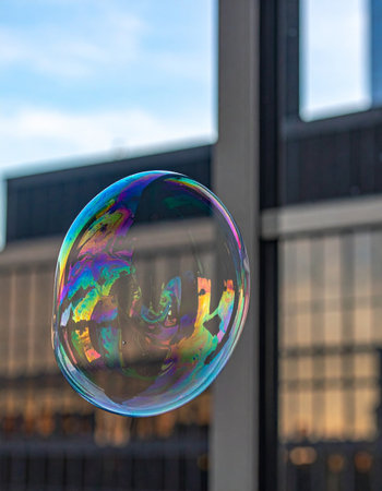 A single, shimmering soap bubble floats in the air, its delicate surface capturing a distorted reflection of the urban landscape and a blue sky.の素材