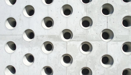 A close-up view of a modern architectural facade reveals a striking pattern of perfectly circular perforations in raw concrete.の素材