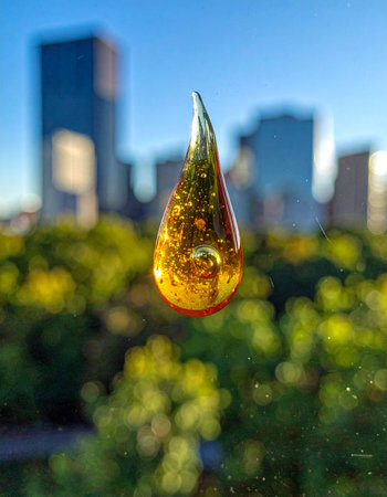 A single, perfect drop of golden liquid hangs suspended in the air, a precious jewel against the blurred backdrop of a modern city and lush greenery.の素材