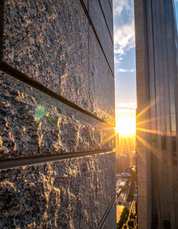 The first golden rays of the morning sun burst between two towering skyscrapers, casting a warm glow on the textured stone facade.の素材