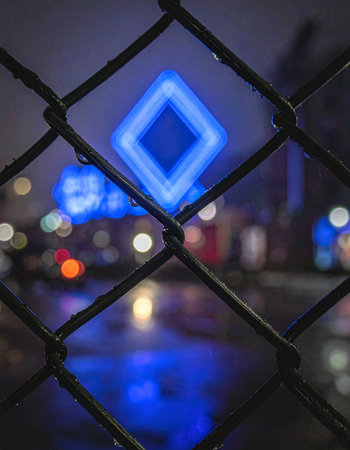 A vibrant blue neon diamond glows mysteriously in the distance on a rainy city night.の素材