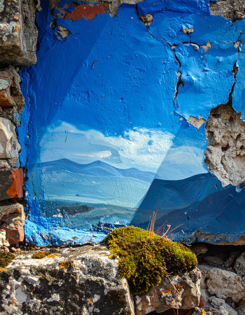 A vibrant blue mountain landscape is painted onto a crumbling, weathered stone wall, creating a surreal window to another world.の素材