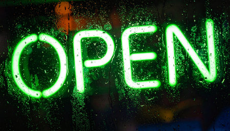 A vibrant green neon 'OPEN' sign glows invitingly through a rain-streaked window at night.の素材