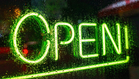 A vibrant green neon 'OPEN' sign glows invitingly through a window streaked with rain.の素材