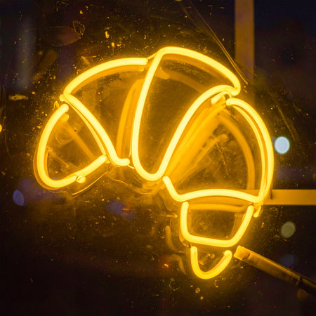 A warm, yellow neon croissant glows invitingly in the dark window of a city bakery, promising fresh pastries and a cozy atmosphere for passersby at night. A modern symbol of a classic treat.の素材