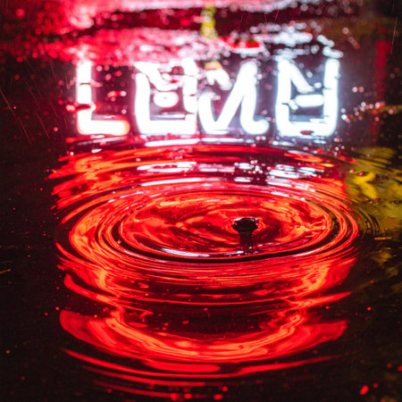 A single drop hits a puddle on a dark city street, sending vibrant red ripples across the reflection of a glowing neon sign.の素材