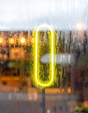 A vibrant yellow neon sign offers a beacon of warmth and light, its glow softly diffused through a window pane streaked with rain.の素材