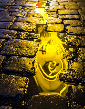 A mysterious golden light with an elegant swirl reflects in a puddle on an ancient, wet cobblestone street at night.の素材