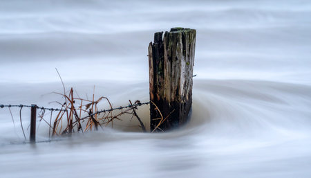 An old, weathered wooden fence post stands resiliently against a smooth, milky current.の素材