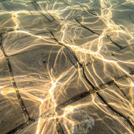 Golden hour sunlight filters through the clear, rippling surface of a pool, casting a mesmerizing dance of light and shadow on the submerged stone floor below.の素材