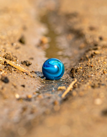 A single, vibrant blue marble, a forgotten relic of a childhood game, rests in a muddy puddle.の素材