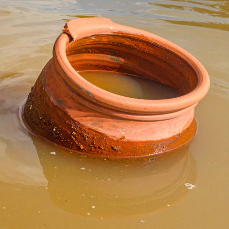A weathered terracotta pipe, a relic of old infrastructure, rests partially submerged in the still, murky water of a flooded area.の素材