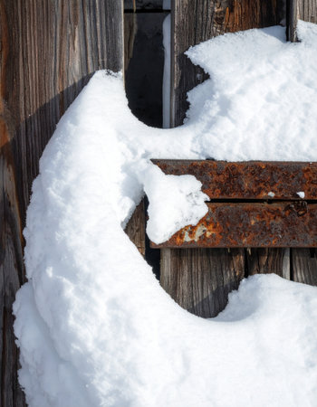 A thick blanket of fresh winter snow clings to a weathered, rusty hinge on an old wooden barn door.の素材