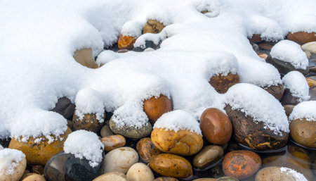 A fresh blanket of pure white snow gently covers a bed of smooth, colorful river stones.の素材