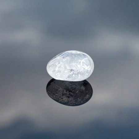 A single, clear crystal, marked by internal fractures, rests in perfect stillness on a reflective plane.の素材