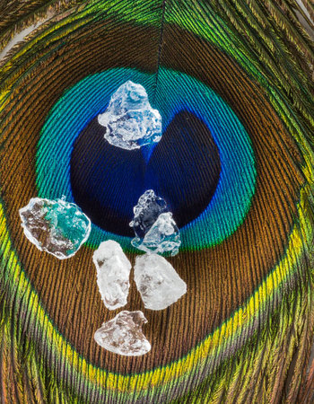 A collection of precious, clear gemstones rests upon the iridescent eye of a peacock feather, creating a luxurious and spiritual still life.の素材