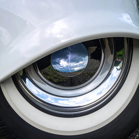 A close-up captures the polished chrome hubcap of a classic car, its mirror-like surface reflecting a distorted, dreamlike image of a blue sky and clouds.の素材