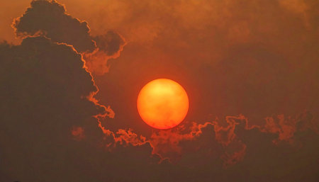 A brilliant orange sun sinks through a thick, moody layer of dark clouds, casting a warm, dramatic glow.の素材