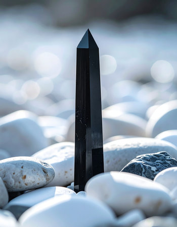 A sleek black crystal obelisk stands tall and powerful amidst a bed of smooth, white stones.の素材