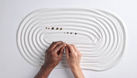 From a top-down perspective, two hands of different skin tones carefully arrange black stones in a white sand zen garden.の素材