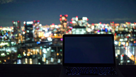 From a high-rise window, a lone laptop is silhouetted against the dazzling lights of the city after dark.の素材