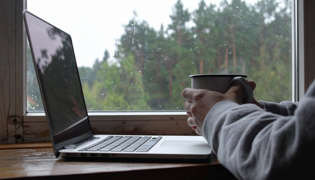A person enjoys a quiet moment, holding a steaming mug of coffee while working on their laptop.の素材