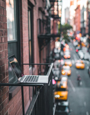 A laptop rests on the railing of a fire escape, offering a unique office view high above a bustling city street.の素材