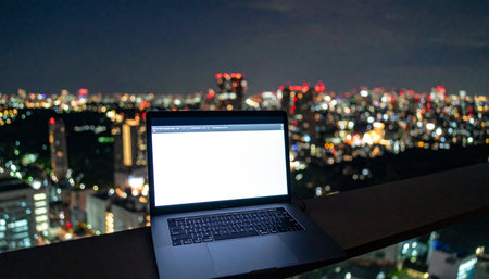 An open laptop with a blank screen sits on a balcony, offering a stunning view of a vibrant city skyline at night.の素材