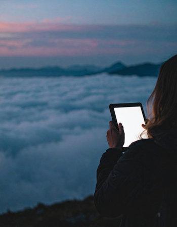 A lone adventurer sits atop a mountain peak at dawn, finding connection and inspiration through a glowing tablet while overlooking a breathtaking sea of clouds.の素材