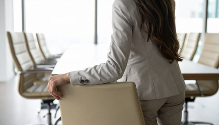 A businesswoman in a tailored suit stands poised by her chair in a sunlit boardroom.の素材