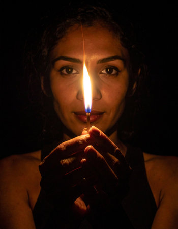 A woman's face emerges from the darkness, her intense gaze fixed on the viewer. She holds a single, steady flame, a symbol of hope, inner strength, and quiet determination in the face of the unknown.の素材