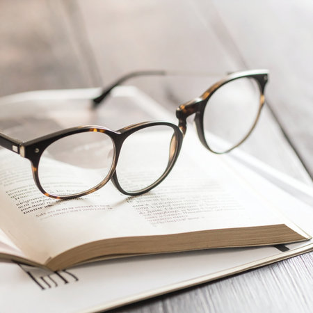 A pair of stylish reading glasses rests on the pages of an open book, marking a pause in a quiet moment of study or leisure.の素材