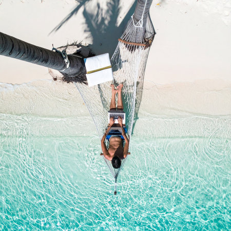An aerial view captures the ultimate work-life balance as a digital nomad works on a laptop while relaxing in a hammock suspended over the crystal-clear turquoise waters of a tropical paradise.の素材
