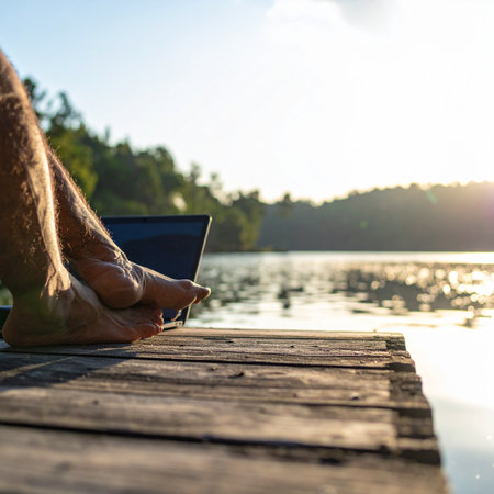 A man enjoys the ultimate work-life balance, sitting on a rustic wooden dock with his laptop.の素材